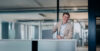 The image shows a man in a beige suit and light blue shirt standing next to his laptop at a desk in a modern office environment.