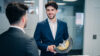 Smiling engineer in a business suit holding an industrial rotor part while discussing technology in a modern office environment.