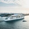 Cruise ship sailing through Hamburg Harbor with container terminals and cranes in the background.